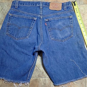 Puede incluir: Pantalones cortos de mezclilla azul con bordes deshilachados y una etiqueta Levi's en el bolsillo trasero.