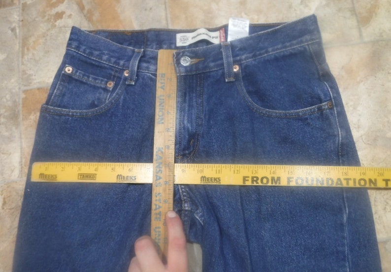 May include: Blue denim jeans with a wooden ruler measuring the waist. The ruler shows measurements in inches.