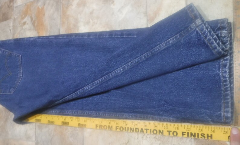 May include: A pair of blue denim jeans folded in half, with a yellow measuring tape placed along the bottom edge. The tape reads "From Foundation to Finish" and has markings in inches.