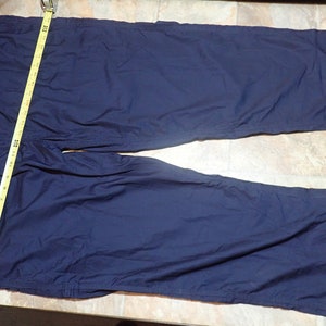 May include: A pair of dark blue pants with a seam down the outside of each leg. The pants are laid flat on a light brown surface.