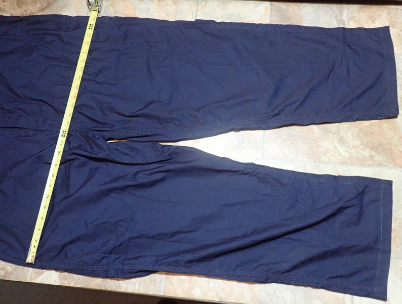 May include: A pair of dark blue pants with a seam down the outside of each leg. The pants are laid flat on a light brown surface.