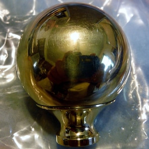 May include: A shiny gold metal sphere with a smooth, reflective surface. The sphere is attached to a round base with a decorative design.