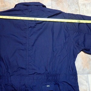 May include: A navy blue short-sleeved jumpsuit with a button-up collar and a drawstring waist. The jumpsuit is made of a lightweight fabric and has a relaxed fit. The measurement across the back is 25 inches.
