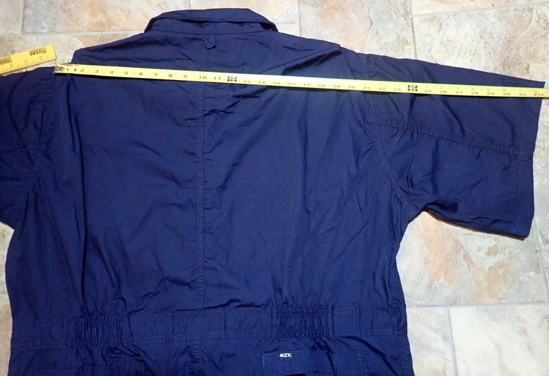 May include: A navy blue short-sleeved jumpsuit with a button-up collar and a drawstring waist. The jumpsuit is made of a lightweight fabric and has a relaxed fit. The measurement across the back is 25 inches.