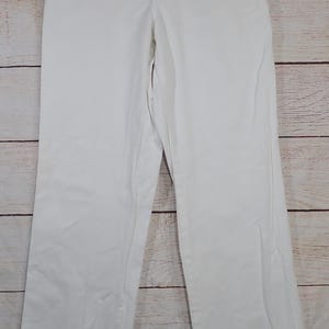 May include: White pants with a straight leg design. The pants have a button and zipper closure, with belt loops and pockets. A tag is attached to the pants. The pants are hanging on a hanger against a light wood background.