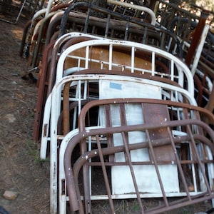 May include: A stack of rusty metal bed frames, some painted white, with a few frames in the background. The frames are stacked in a pile, with the top frame being the most visible. The frames are all different sizes and shapes, but they are all made of metal.