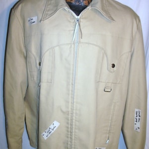 May include: A beige zip-up jacket with a white zipper and two pockets on the front. The jacket has handwritten notes on it that say "I'm Stan", "Google Maps", "Am still spots", "I had plenty of spooky soup", and "I'm Stan".