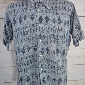 May include: A short-sleeved button-down shirt with a geometric pattern in shades of gray and white. The shirt features a classic collar and button closure. The pattern includes diamond and triangle shapes.