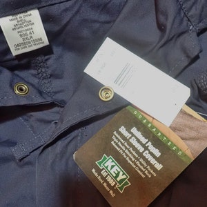 May include: A navy blue short-sleeve coverall with a white tag that reads "Unlined Poplin Short Sleeve Coverall" and "Guaranteed". The tag also includes the brand name "KEY Est 1908" and the text "Works Hard. Wears Well".