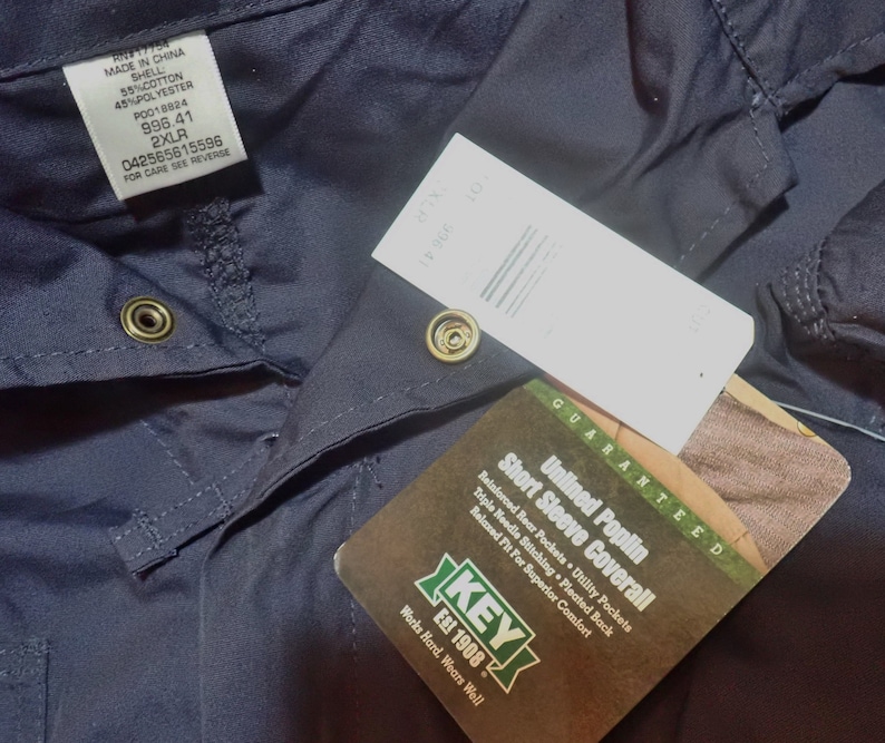 May include: A navy blue short-sleeve coverall with a white tag that reads "Unlined Poplin Short Sleeve Coverall" and "Guaranteed". The tag also includes the brand name "KEY Est 1908" and the text "Works Hard. Wears Well".