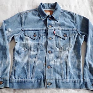 May include: Light blue denim jacket with a bleached pattern. The jacket has a classic collar, button-down front, two chest pockets, and buttoned cuffs. A small red tag is visible on the left chest pocket.