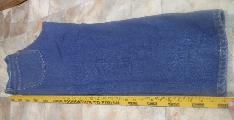 May include: A pair of blue denim jeans laid flat. The jeans have a single back pocket and a yellow measuring tape is placed below the jeans.