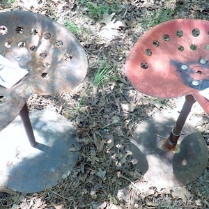 Könnte beinhalten: Zwei alte Traktorsitze, einer rostbraun und der andere rot, jeweils mit einem Metallsockel und einer zentralen Stütze. Die Sitze haben mehrere kreisförmige Löcher. Jeder Sitz hat ein Blatt Papier mit einem Buchstaben.