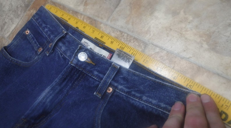 May include: A close-up of the waistband of a pair of blue denim jeans. The jeans have a button fly and a Levi's brand label.