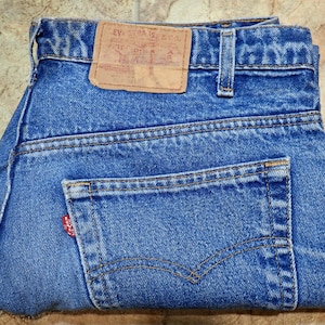 Könnte beinhalten: Blaue Jeans aus Denim mit einem hellbraunen Lederaufnäher am hinteren Bund, der den Text Levi Strauss & Co. trägt. Die Jeans verfügt über eine Gesäßtasche mit einem kleinen roten Etikett an der Seite, Gürtelschlaufen und sichtbare orangefarbene Nähte.