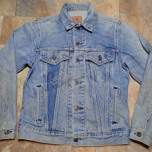 May include: Light blue denim jacket with button closures, two chest pockets, and two side pockets. The jacket has a classic collar and buttoned cuffs. The denim has a faded, worn look, suggesting it's a vintage or well-loved piece.