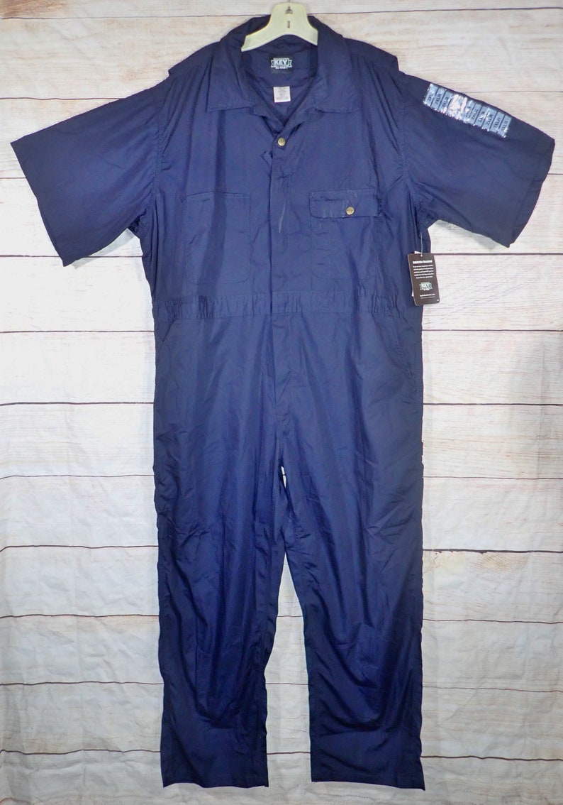 May include: A navy blue short-sleeved jumpsuit with a button-up front and a single chest pocket. The jumpsuit has a brand label that reads "KAVU" on the left side of the chest.