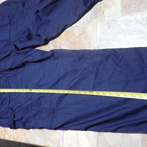 May include: A dark blue work jumpsuit with multiple pockets and a drawstring waist. The jumpsuit is laid flat on a light brown surface with a yellow measuring tape.