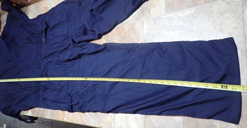 May include: A dark blue work jumpsuit with multiple pockets and a drawstring waist. The jumpsuit is laid flat on a light brown surface with a yellow measuring tape.