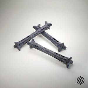 Può includere: Tre tasselli decorativi grigi e stilizzati. Ogni tassello ha un design strutturato e organico con una serie di fori lungo l'asta e un'estremità a forma di stella. I tasselli sono disposti su uno sfondo bianco.