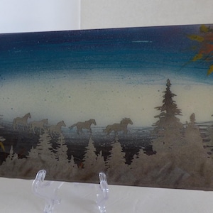 May include: Rectangular art piece depicting a landscape with running horses and silhouetted pine trees. The sky transitions from dark blue to light blue, with a yellow and orange burst in the upper right corner. The artwork is displayed on a clear stand.