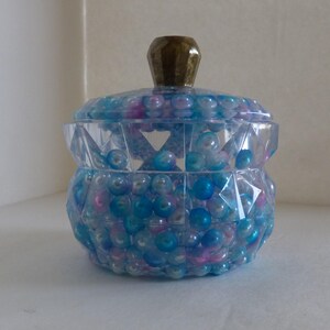 May include: A clear, faceted jar filled with blue, pink, and clear beads. The jar has a matching lid and a gold-colored knob. The jar is a decorative item.