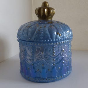 Könnte beinhalten: Ein blaues Glasgefäß mit einem dekorativen kronenförmigen Deckel. Das Gefäß hat ein verziertes Design mit goldenen Akzenten. Das Gefäß hat eine hellblaue Farbe mit Perlmutt-Finish. Der Deckel ist goldfarben.