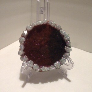 May include: A polished, round geode with a deep burgundy center and a sparkling, crystal-like border. The geode is displayed on a clear plastic stand, showcasing its natural beauty and unique texture. The geode is a decorative item.