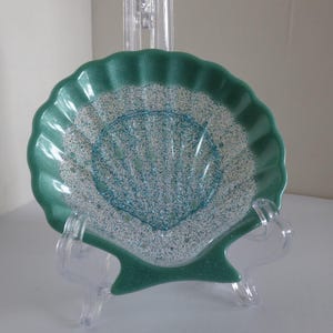 May include: A teal seashell-shaped decorative plate with a glittery, textured surface. The plate is displayed on a clear plastic stand. The plate's rim has a scalloped edge. The plate is a decorative item.