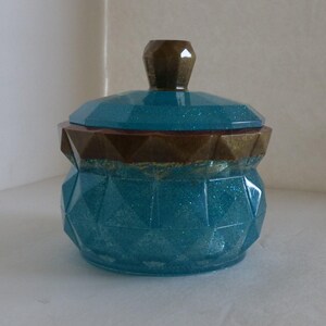 May include: A teal and gold decorative jar with a lid. The jar has a geometric design with a teal glitter finish. The lid is teal with a gold rim and a gold knob. The jar is likely used for storage or decoration.