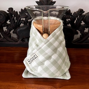 May include: A glass coffee carafe with a wooden collar, wrapped in a quilted sage green and white gingham fabric cozy. The fabric wrap features a small white rectangular tag with the text cozi printed in black, secured by a wooden bead.