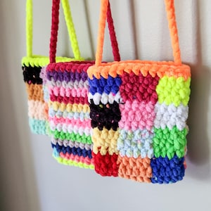 以下が含まれることがあります： ニュートラルな壁に掛けられた、鮮やかな色の手作りのかぎ針編みバッグが3つ。バッグは、ピンク、青、黄色、オレンジなど、さまざまな色のパッチワークデザインが特徴です。バッグのストラップと上端は、対照的な明るい色です。