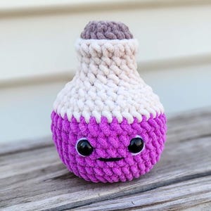 Könnte beinhalten: Eine handgefertigte, gehäkelte Zaubertrankflasche in Lila, Weiß und Braun. Die Flasche hat ein lächelndes Gesicht mit schwarzen Augen und einem aufgestickten Mund. Das Plüschtier sitzt auf einer Holzoberfläche.