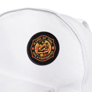 Könnte beinhalten: Schwarz-weißer Patch mit einem Feuerwehrmann, der eine andere Person umarmt. Der Patch trägt den Text "Im Brandfall einen Feuerwehrmann umarmen" in gelber und orangefarbener Schrift.