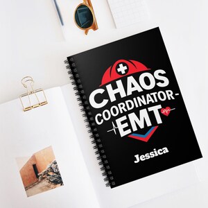 Op de afbeelding: Een zwarte spiraalnotitieblok met een wit ontwerp dat "Chaos Coordinator - EMT" zegt met een rood kruis en een rood hart met een hartslaggrafiek. De naam "Jessica" staat onderaan het notitieblok gedrukt.
