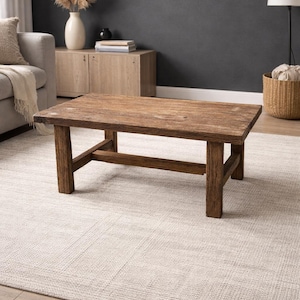Könnte beinhalten: Ein rechteckiger Couchtisch aus Holz mit natürlicher Oberfläche. Der Tisch hat ein schlichtes, robustes Design mit vier Beinen und einem unteren Stützbalken. Er steht auf einem hellen, gewebten Teppich in einem Wohnzimmer.