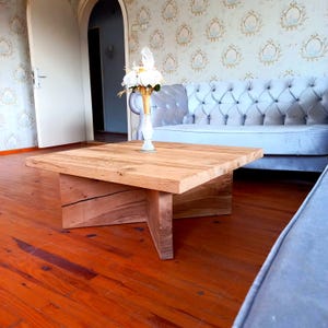 Könnte beinhalten: Ein hellbrauner Couchtisch aus massivem Holz mit quadratischer Oberseite und einzigartigem Sockeldesign. Der Tisch ist ca. 91 cm lang und breit und 41 cm hoch. Auf dem Tisch steht eine Vase mit weißen Blumen.