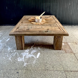 May include: A rustic wooden coffee table with a natural wood finish. The table has a square top and four legs. A small silver bowl with a wooden spoon sits on a woven mat in the center of the table.