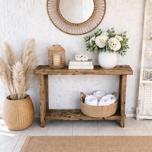 Puede incluir: Una consola de madera rústica con una cesta tejida, toallas blancas y un jarrón con flores blancas. Un espejo redondo tejido cuelga sobre la mesa. Una cesta tejida con hierba seca alta y una alfombra tejida completan la decoración.