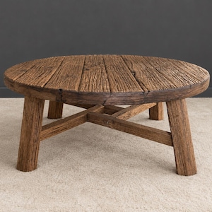Könnte beinhalten: Ein runder Couchtisch aus verwittertem braunem Holz mit sichtbarer Maserung. Die Tischplatte besteht aus mehreren zusammengefügten Dielen und wird von vier dicken, geraden Beinen getragen, die durch eine kreuzförmige Verstrebung verbunden sind.