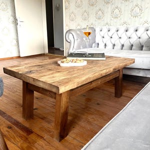 Könnte beinhalten: Ein rustikaler Holz-Couchtisch mit einer natürlichen Holzoptik. Der Tisch hat eine rechteckige Tischplatte und vier robuste Beine. Der Tisch steht auf einem Hartholzboden in einem Wohnzimmer.