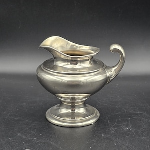 May include: A silver-coloured creamer with a curved handle and a spout. The creamer has a bulbous body and sits on a pedestal base. The handle curves gracefully, and the spout is designed for easy pouring. The creamer is made of a shiny, reflective metal.