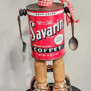 May include: A whimsical doll made from a vintage Savarin coffee can, with a black face, red and white checkered scarf, and wooden legs. The doll is wearing copper-colored shoes and holding a fork and spoon.
