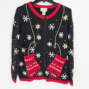 May include: Black zip-up sweater with red trim and accents. The sweater features white and gold snowflakes, red mittens, and a gold thread design. A festive winter holiday theme.