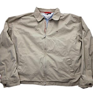 May include: A tan zip-up jacket with a collared neckline. The jacket has a small logo on the chest and a red and blue striped lining. It features two front pockets and buttoned cuffs. The jacket is a neutral color.