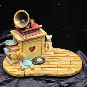 May include: A whimsical figurine featuring a vintage gramophone with a golden horn atop a stack of books. A small clown and a cat sit nearby. The base is a wooden platform with a heart on the gramophone.