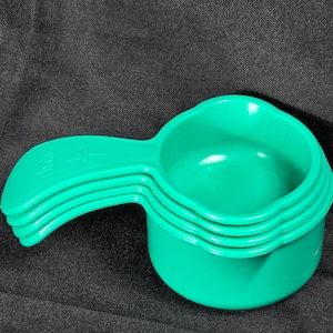 Puede incluir: Un conjunto de cuatro tazas medidoras verdes apiladas. La taza superior tiene la inscripción "cup" y "1/3" en el mango. Estas tazas de plástico son de color verde brillante y se utilizan para medir ingredientes en la cocina.