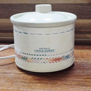 May include: Cream-colored Rival Little Dipper electric stoneware server with a lid. The server has a decorative band of colorful arrows and geometric shapes. The text "RIVAL LITTLE DIPPER ELECTRIC STONEWARE SERVER" is printed on the front.