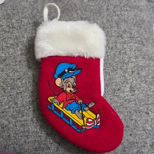 May include: Red Christmas stocking with a white faux fur cuff and a loop for hanging. The stocking features a cartoon mouse wearing a blue hat and red shirt, riding a yellow sled. The mouse is smiling.