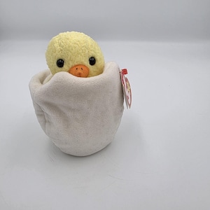 May include: A soft toy chick with a yellow body, black eyes, and an orange beak, emerging from a white eggshell. The chick is made of a textured material. A red tag is attached to the eggshell. The toy is designed to look like a baby chick hatching from an egg.